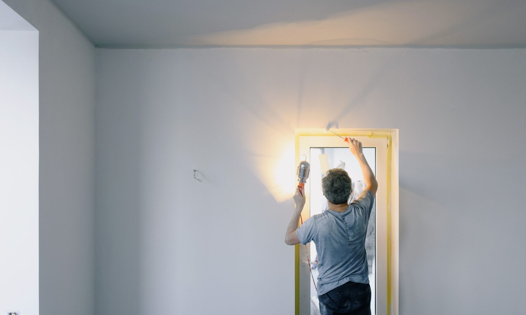 Mann streicht mit einer Farbrolle eine Innenwand neben einem Fenster