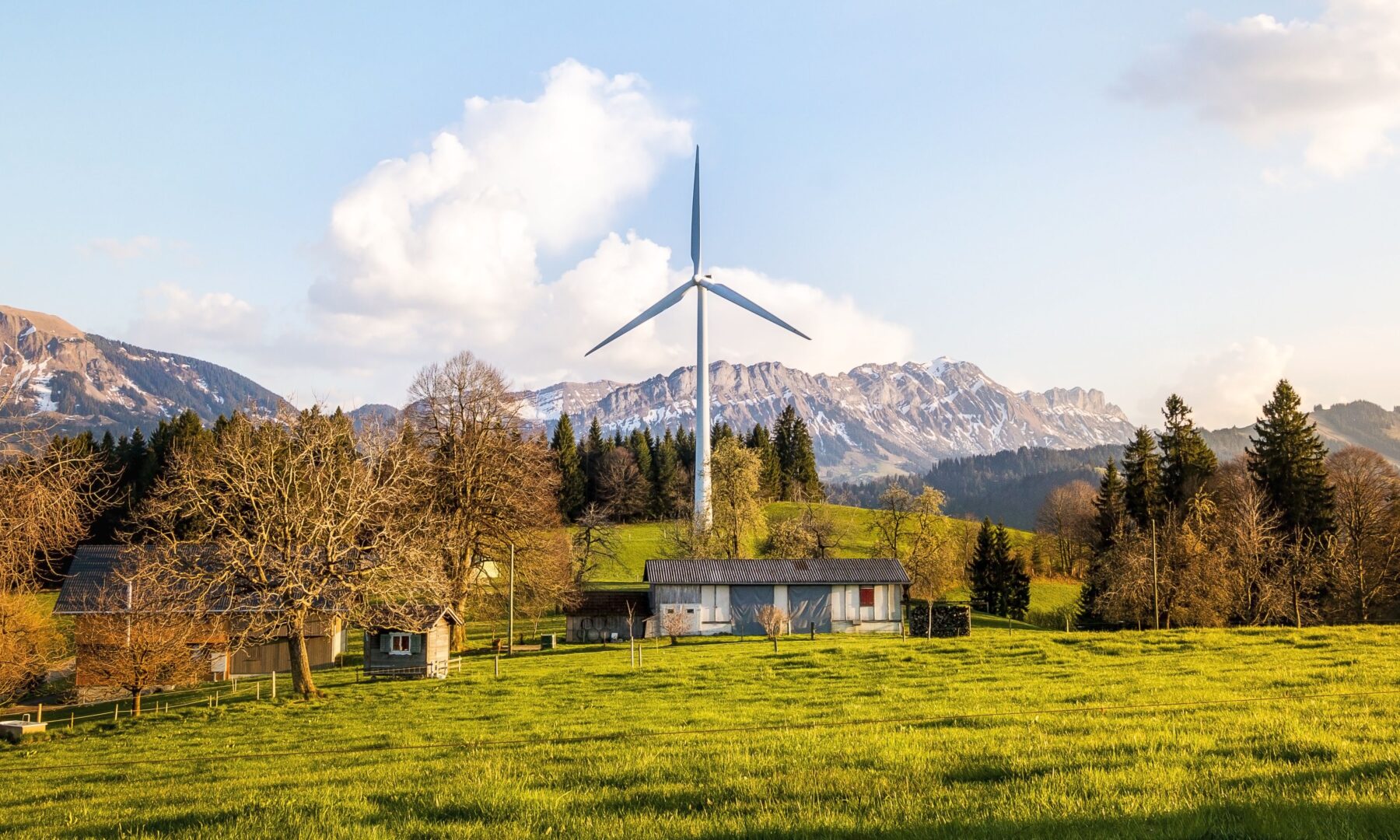 Einzelne Windenergieanlage auf einer grünen Wiese neben landwirtschaftlichen Gebäuden, im Hintergrund eine Bergkette.