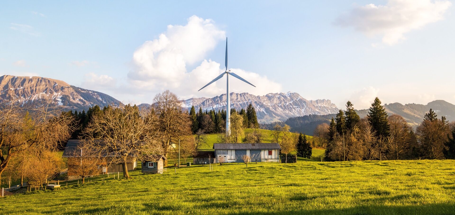 Einzelne Windenergieanlage auf einer grünen Wiese neben landwirtschaftlichen Gebäuden, im Hintergrund eine Bergkette.