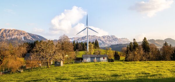 Einzelne Windenergieanlage auf einer grünen Wiese neben landwirtschaftlichen Gebäuden, im Hintergrund eine Bergkette.