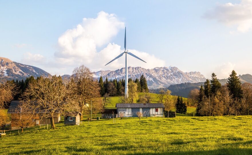 Einzelne Windenergieanlage auf einer grünen Wiese neben landwirtschaftlichen Gebäuden, im Hintergrund eine Bergkette.